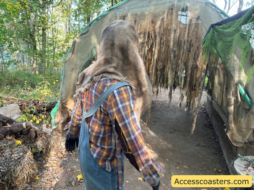 A person in a checked shirt and creepy burlap mask walks through a dimly lit scare zone tunnel lined with straw and hanging decorations, with branches and foliage around the entrance.