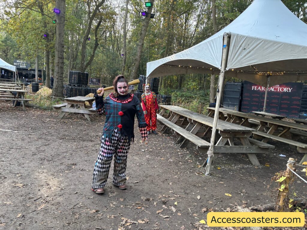 The clown actor poses playfully on a woodland path next to picnic benches and a white tent, with the "Fear Factory" sign in the background.