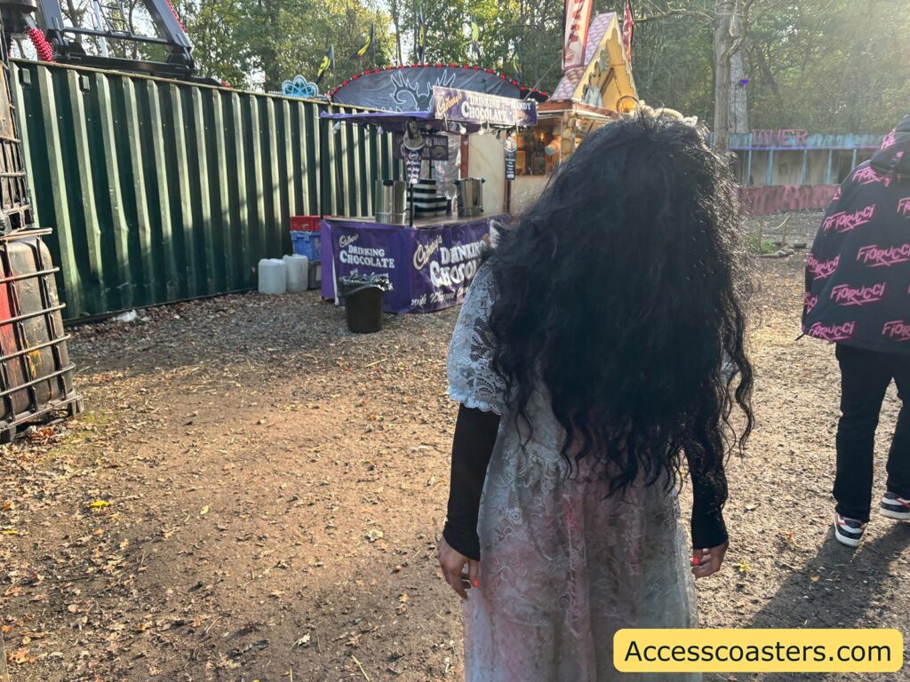 A spooky actor with long, messy dark hair and a tattered dress stands near green shipping containers marked "Fear Factory."