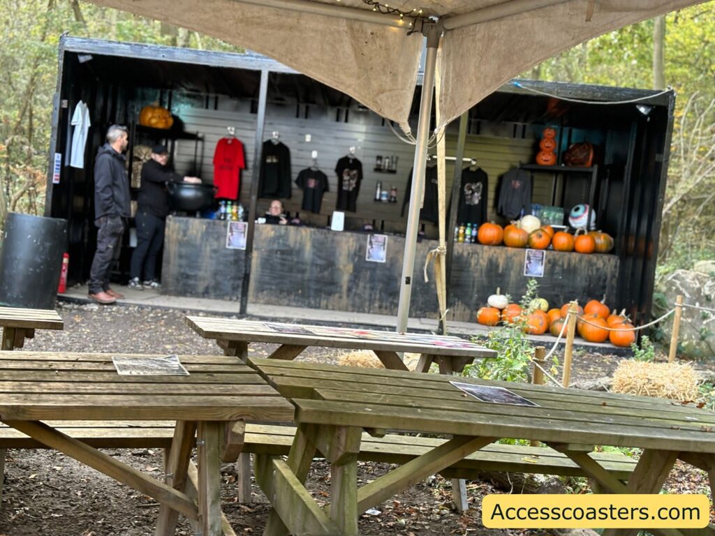 An outdoor stall under a canopy with pumpkins lined up for sale on shelves, a few people attending the stall, and wooden tables in the foreground for pumpkin carving.