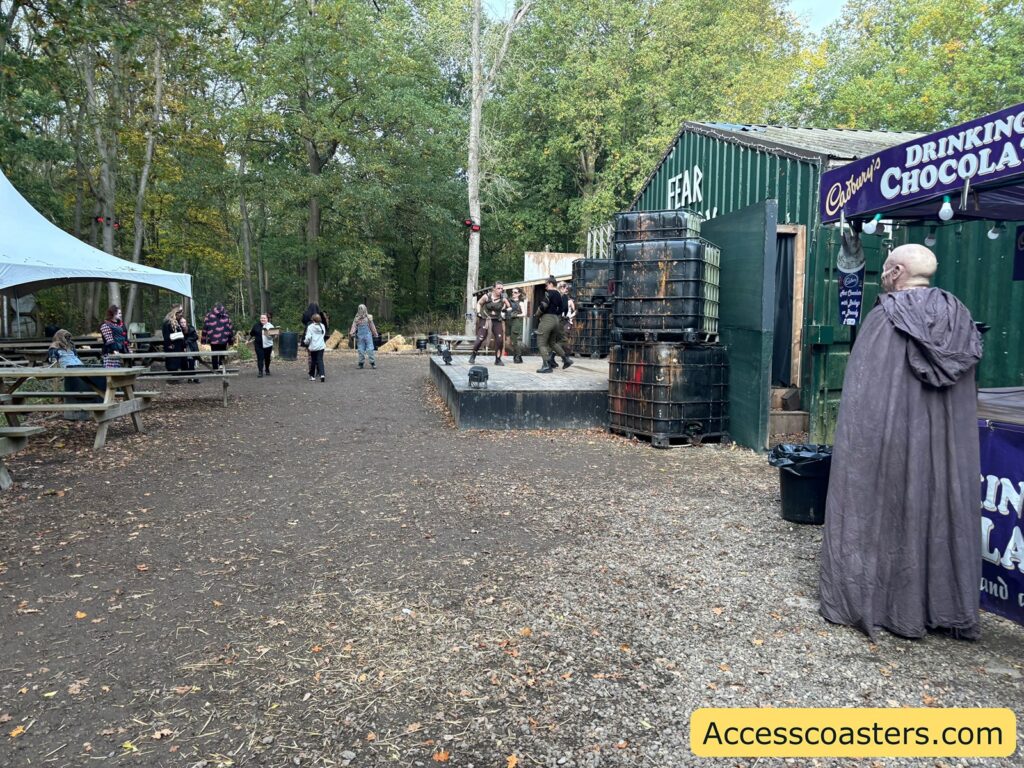 Event guests walk and sit on picnic benches near a wooded area as a person in a long, hooded cloak stands by a drinks stand