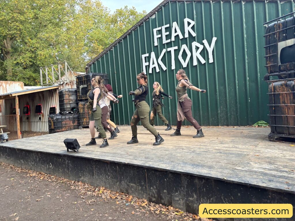 A group of performers in brown costumes dance on an outdoor wooden stage in front of a green building with a large "FEAR FACTORY" sign 