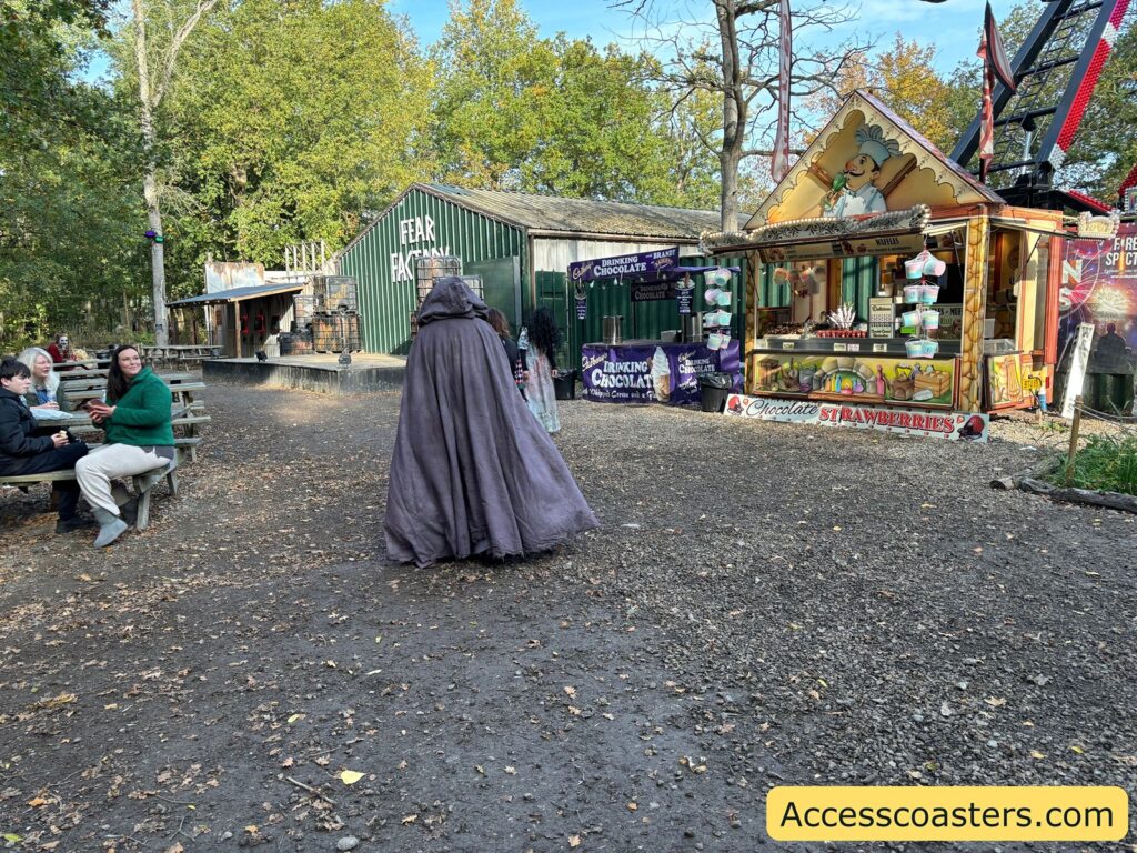 An actor in a long cloak walking through a woodland area with picnic benches, food stalls, and people sitting and eating at Spookfest.
