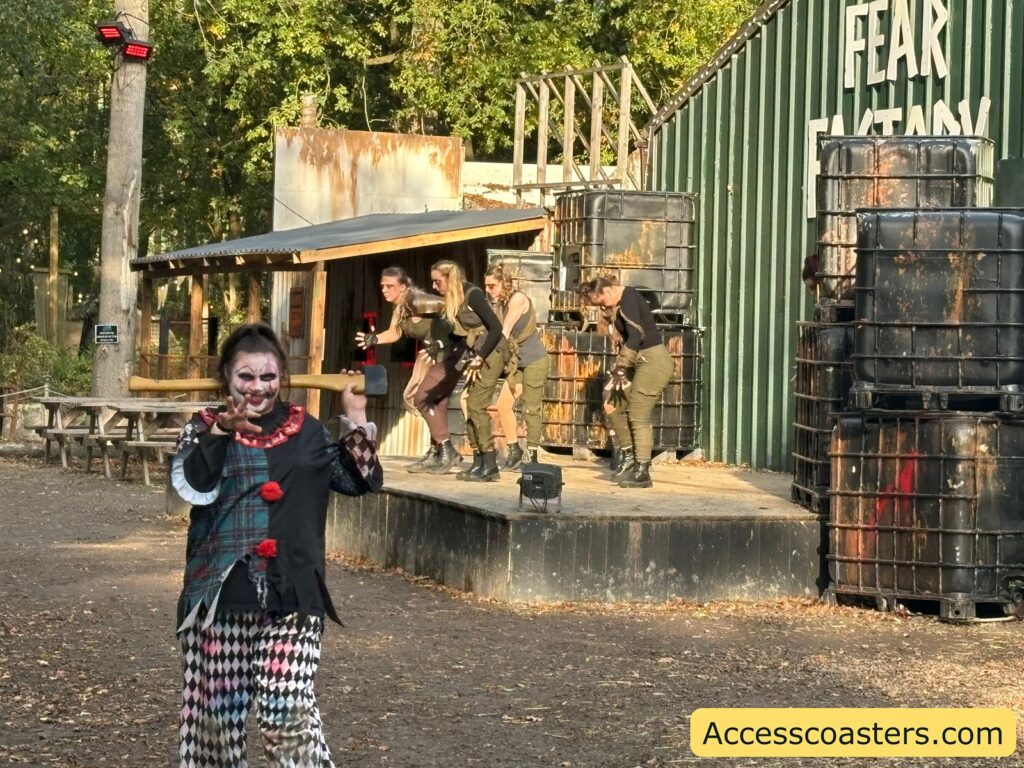an actor dressed as a creepy clown, with a painted face and checkered costume, holding up their arms and standing in front of a stage with Halloween decorations.