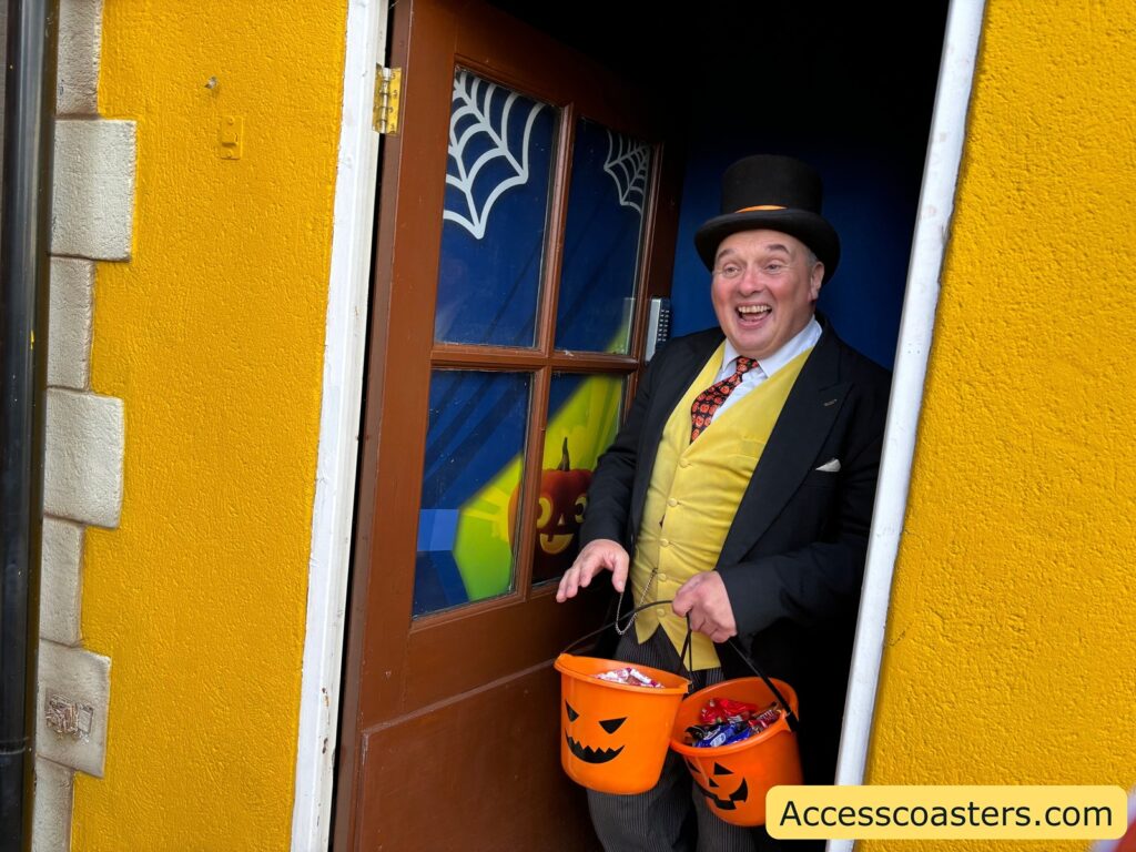 Sir Topham Hat character, wearing a black hat and yellow vest, stands in the doorway of the yellow house at Thomas Land, smiling and holding a pumpkin bucket filled with treats for trick-or-treaters.