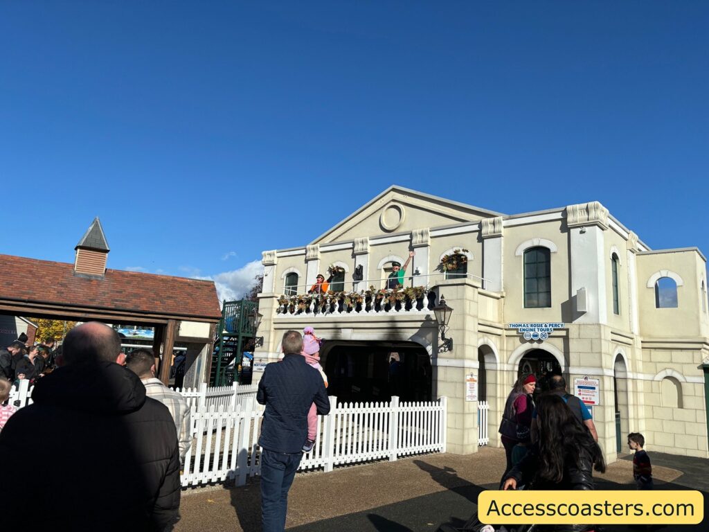 A group of visitors stand below the white Thomas Land building, with children and adults looking up at the balcony where characters perform a show. 