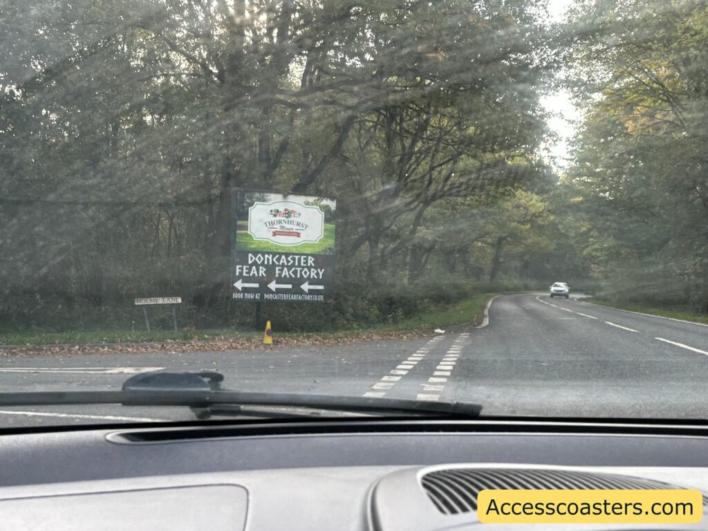 Yellow road sign reading “FEAR FACTORY” with an arrow, positioned beside a narrow driveway and lined with small cones, directing cars towards the event parking area.
