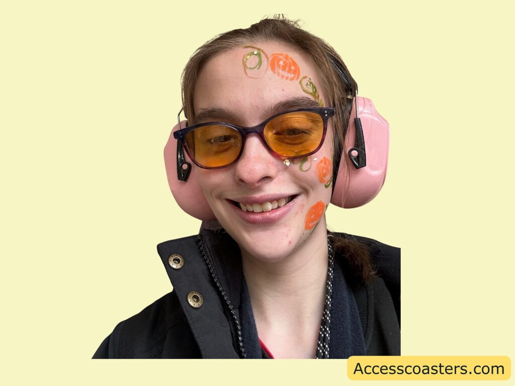 image of Iona with orange tinted glasses and pink earmuffs, showing Halloween-themed face paint featuring pumpkins and swirls.
