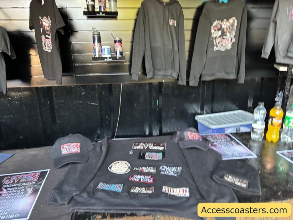 A table at the merchandise stall displaying Fear Factory branded items such as hoodies, t-shirts, beanies, and accessories, with more clothing items hanging on racks in the background.