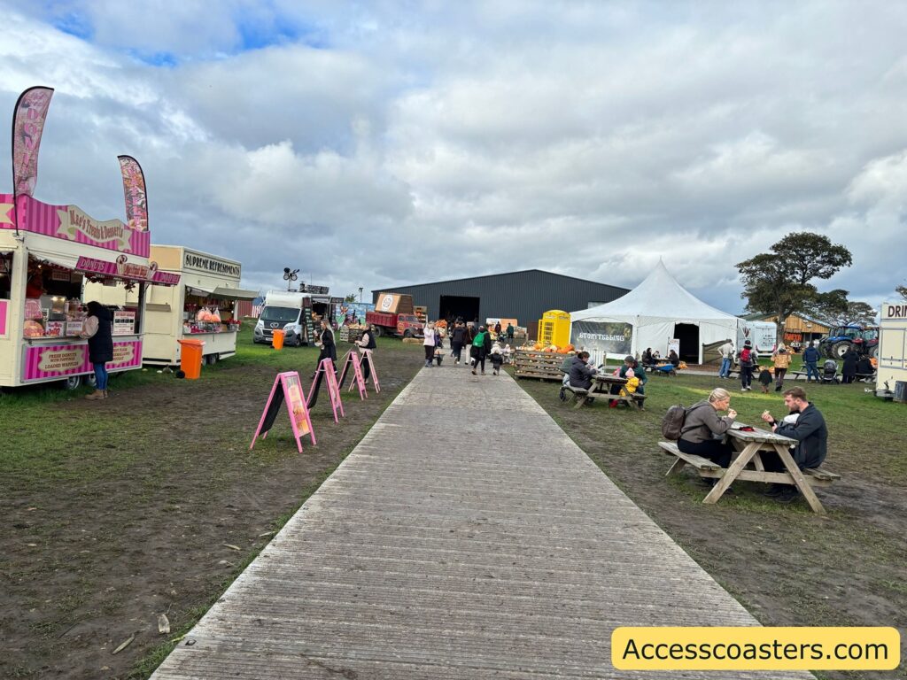 Photo of a boardwalk-style path laid over grass at an outdoor festival, with food stalls, picnic tables, and people along the walkway  there is muddy ground around the path