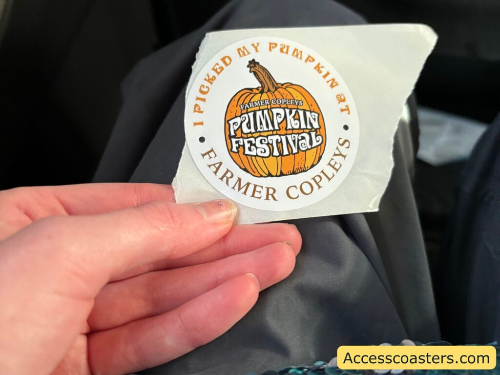  hand holding a round sticker that reads "Proud By Popular Demand Farmer Copleys Pumpkin Festival."