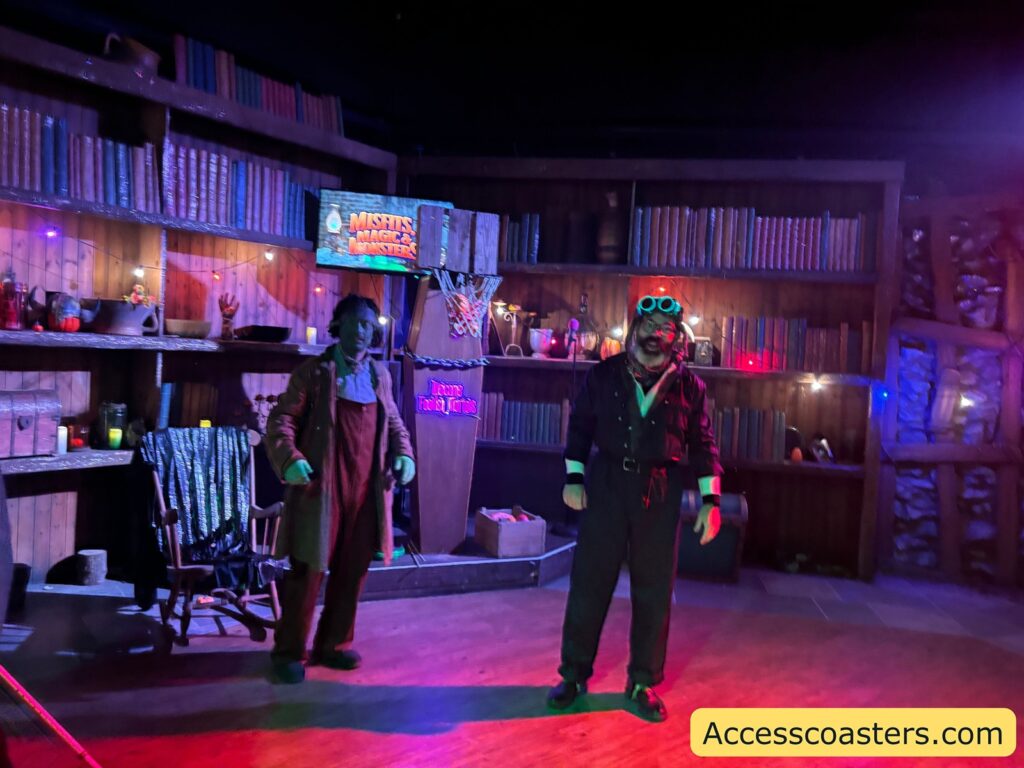 A dimly lit room decorated like a library, with shelves full of books and Halloween props. Two people are present; one is dressed as Frankenstein, welcoming guests as part of a Halloween-themed activity.
