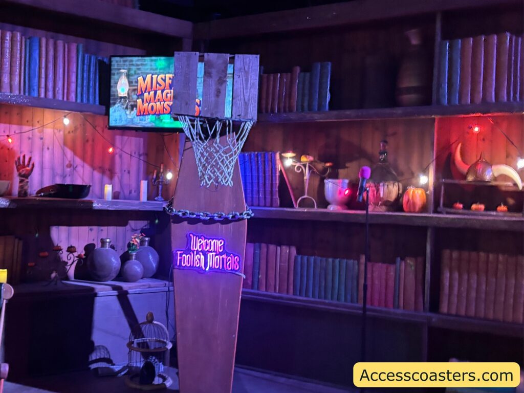 A close view of the themed basketball game setup, showing a basketball hoop with a monster-themed backboard and Halloween objects arranged below it, set against shelves of colorful books and spooky decorations.