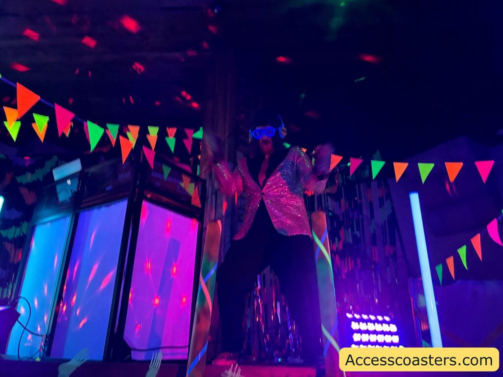 The DJ standing on stage under UV lights, surrounded by bright bunting, as music and light beams fill the room