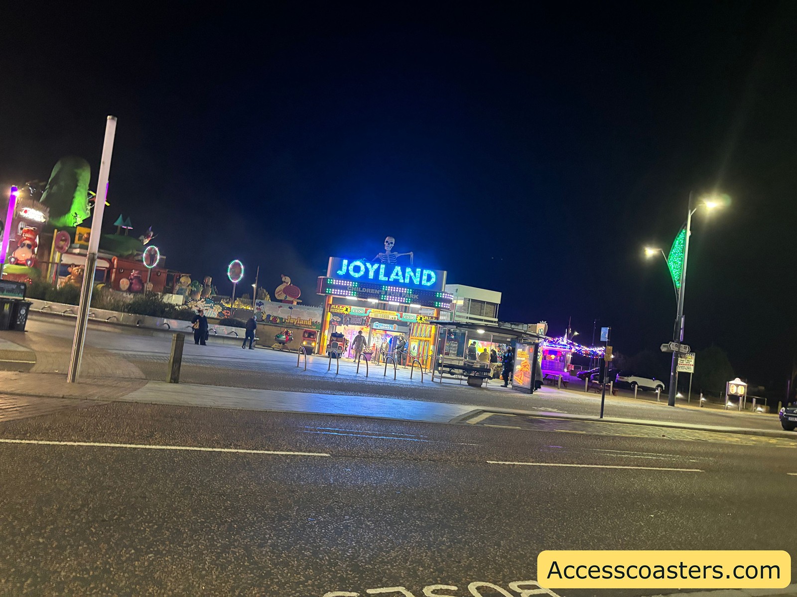 Joyland amusement park entrance at night, brightly lit with a large sign and colorful lights, viewed from across the street.