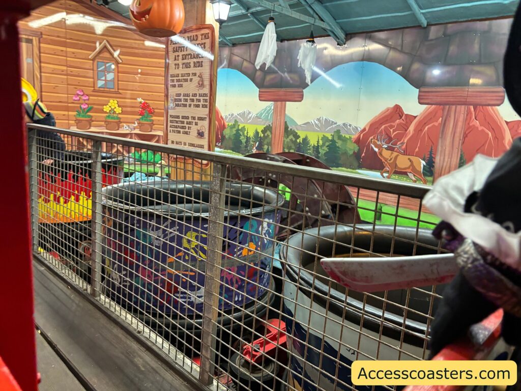 Side view of spinning tubs at the Tyrolean Tub Twist ride, surrounded by safety railings and a vibrant, cartoon-style backdrop with mountains and flowers.
