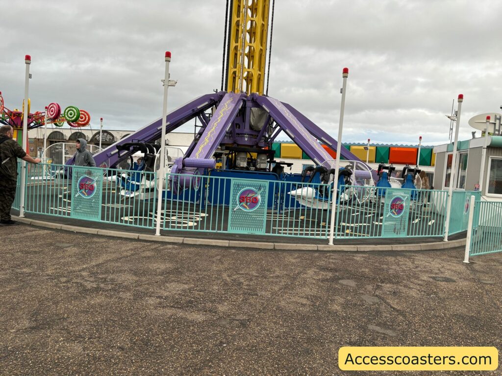 View of the Lightning 360 ride entrance and queue area, surrounded by a light blue fence, with the ride structure behind