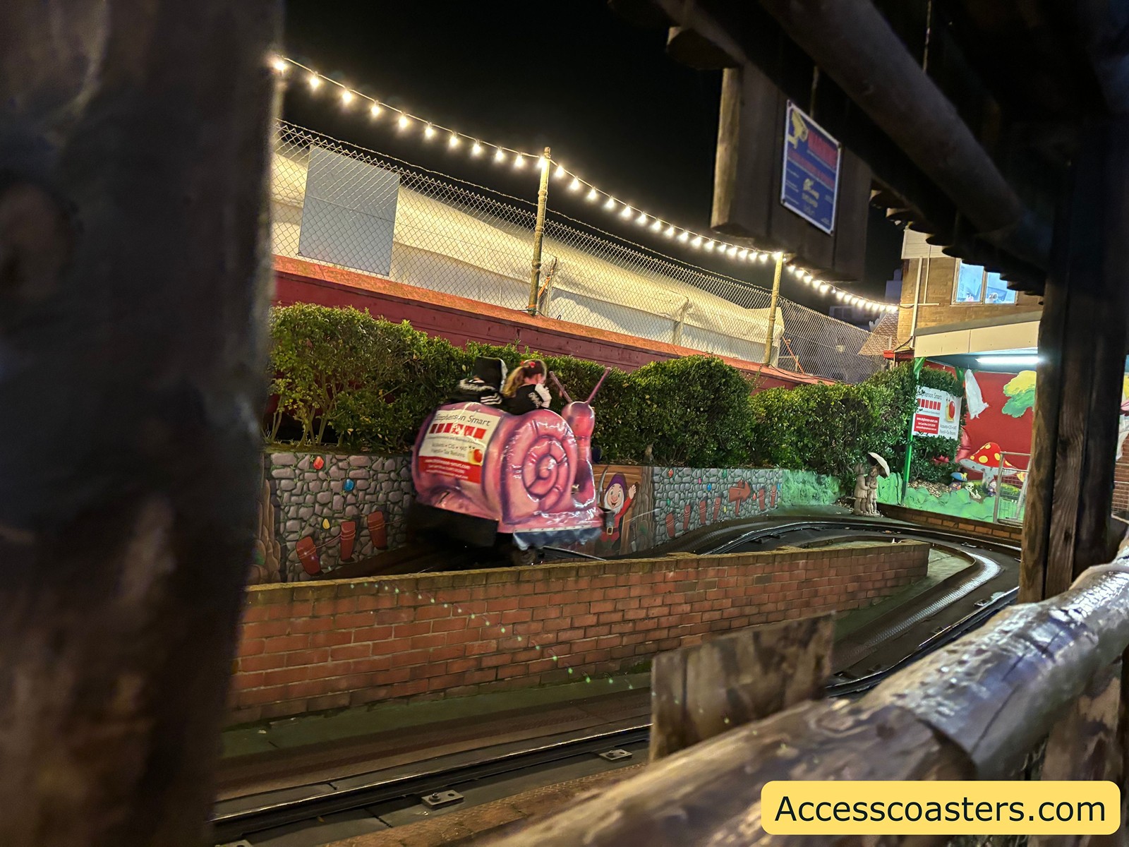 Close-up of a snail-themed ride cart with a passenger, moving on a winding track with brick walls and plants, illuminated by warm string lights at night.