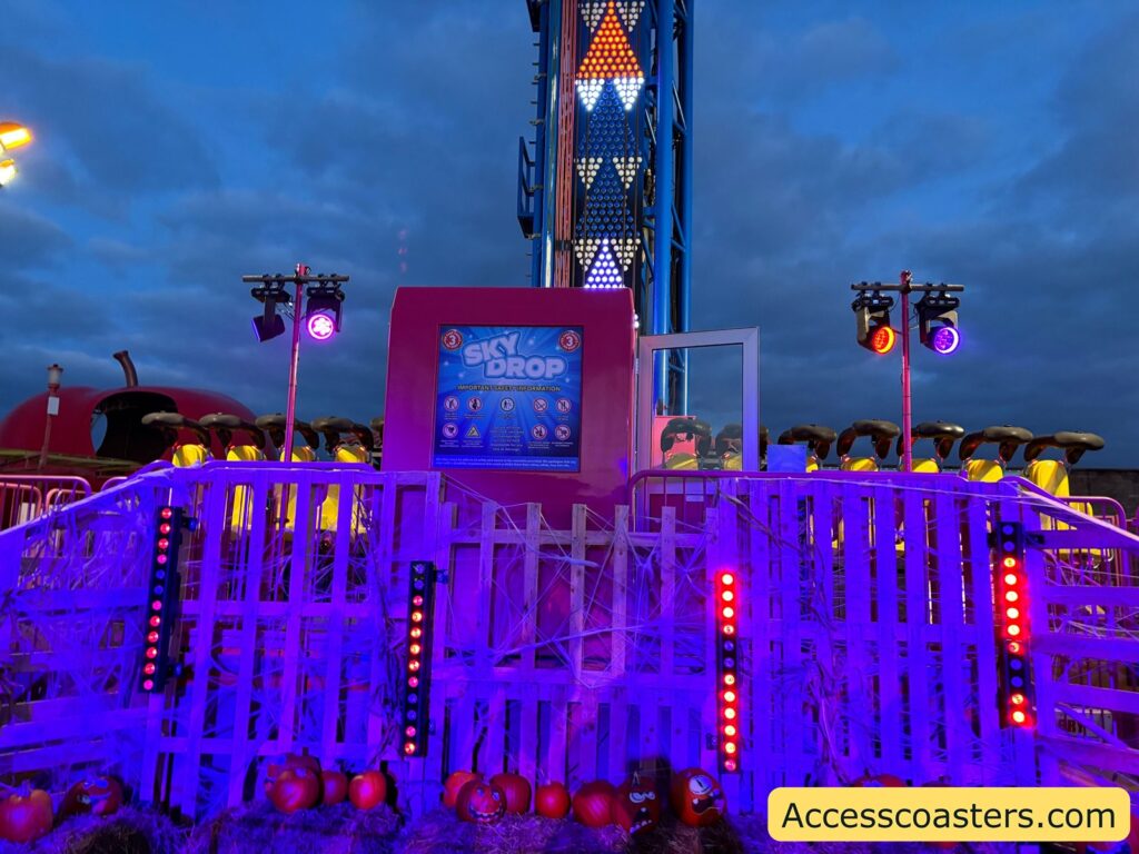  The Sky Drop ride platform with purple fencing, bright lights, and the ride tower in the background, showing how guests board the drop tower