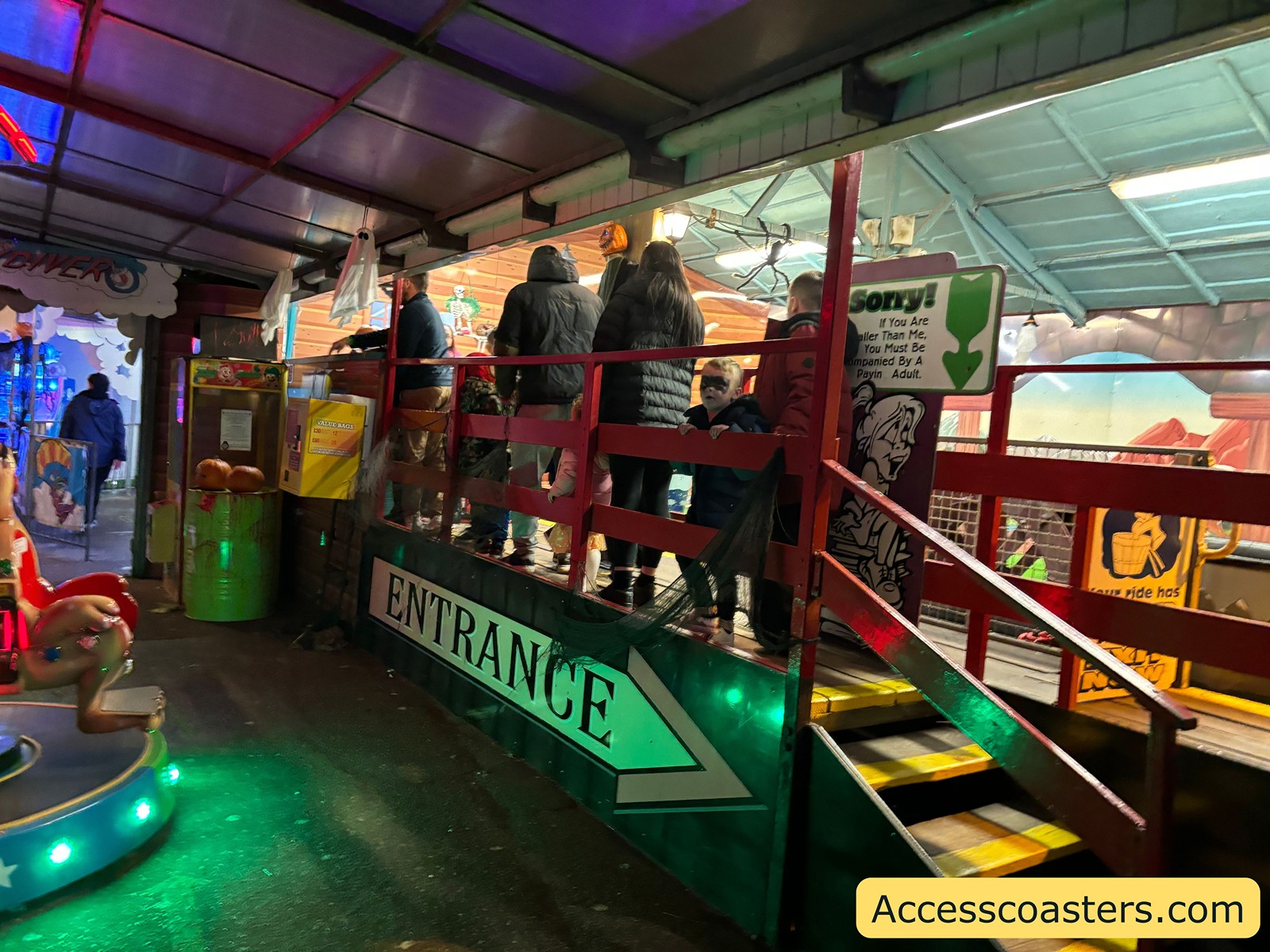 People wait to board the Tyrolean Tub Twist ride at Joyland, standing on a raised platform by an entrance sign, with colorful lights and ride decorations visible.