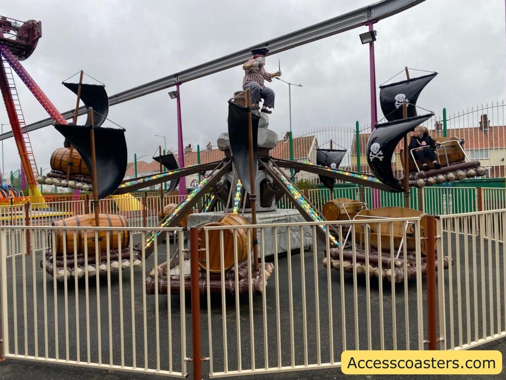 children’s amusement ride shaped like small rafts moves in a circle, with each raft attached to a long arm. The ride is surrounded by a white fence, and there are people riding in the rafts.