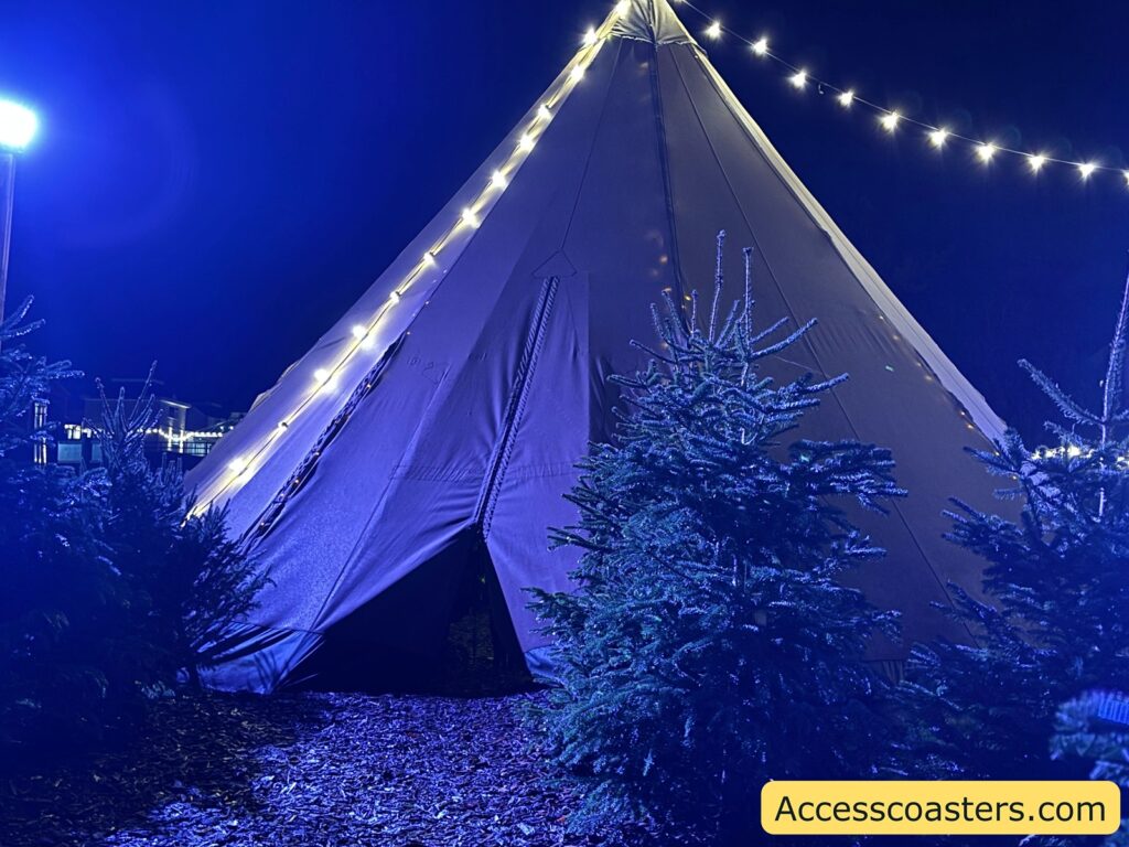  A large, illuminated tent at night, decorated with string lights and surrounded by Christmas trees, marking the entrance to the pranking workshop.