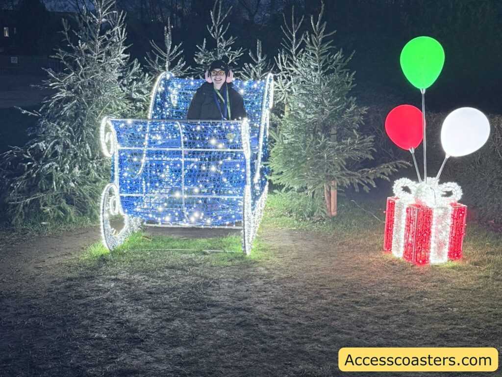 Blue Christmas light display shaped like a sleigh, with large colorful present props and balloon decorations beside it.