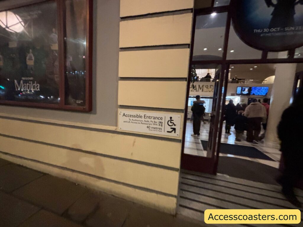 Exterior wall of the theatre with a sign indicating the accessible entrance and a glass door to the right where people are entering the building.