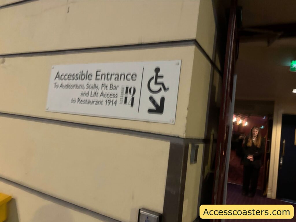 Close‑up of an “Accessible Entrance” sign with wheelchair symbol and arrow mounted on a cream wall beside a doorway inside the theatre.