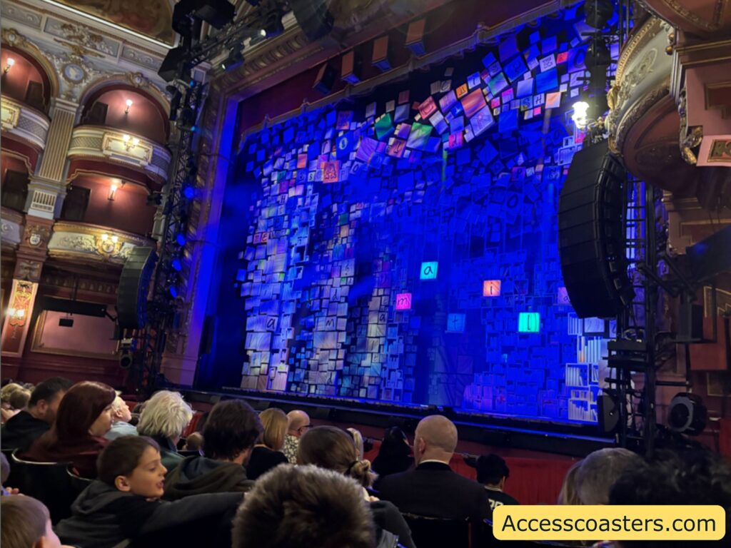 View from the stalls inside a grand theatre, showing the backs of audience members and a brightly lit stage with a colourful set, surrounded by ornate balconies.