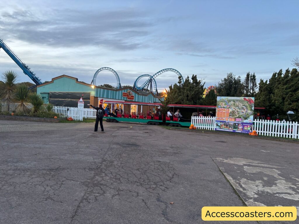 Broad view showing the park entrance gate, a few visitors, and roller coaster tracks visible in the background.
