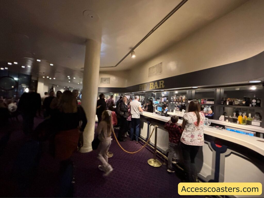 Long theatre bar with people queuing behind a rope barrier, bottles and drinks displayed along the counter, illustrating the accessible bar area available before and during the interval.