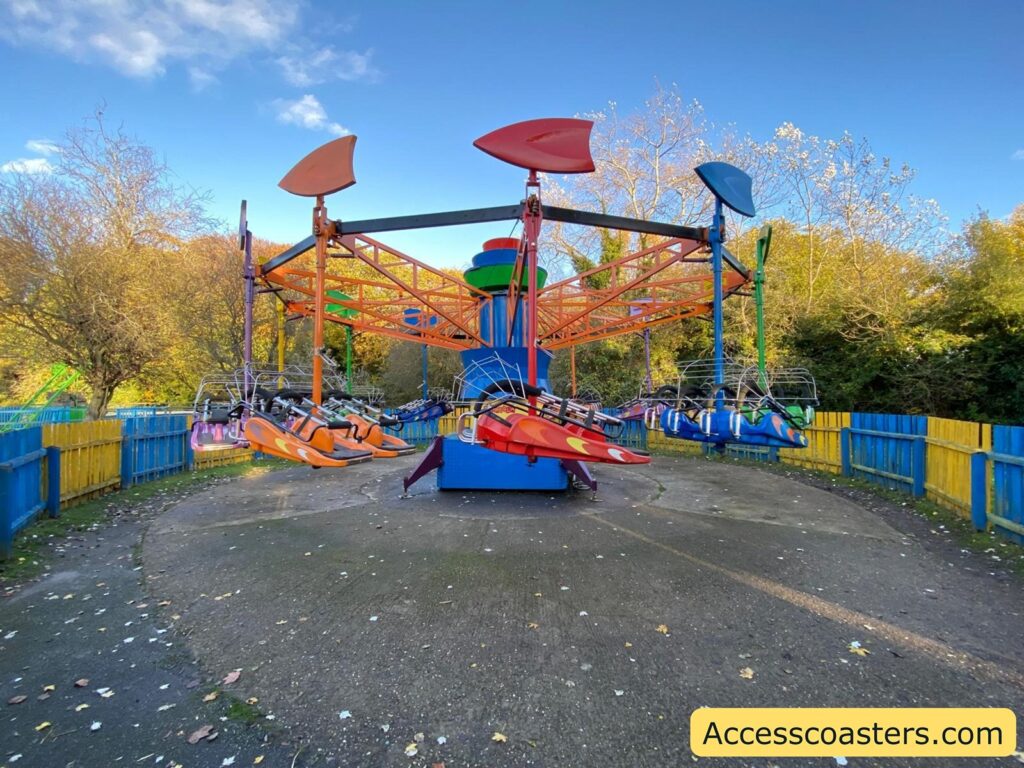 Wide view of the Kite Flyer family ride, showing the ride’s large circular structure and orange ride vehicles surrounded by a blue and yellow fence.