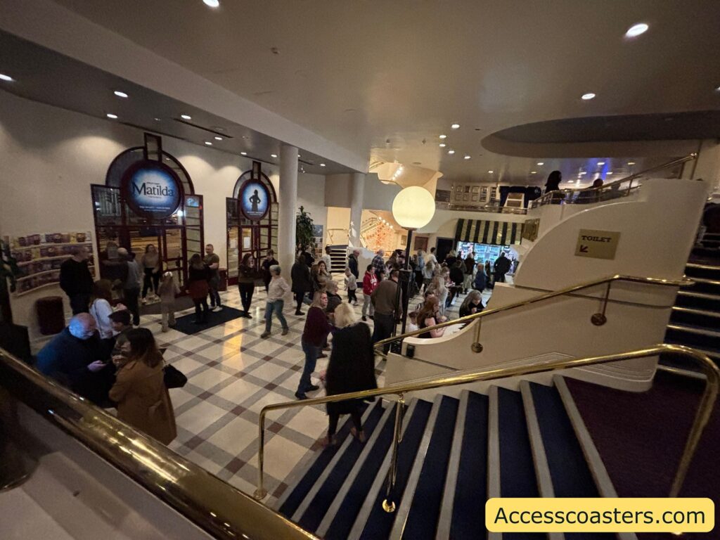 Busy theatre foyer with people moving around, a wide staircase with blue steps and gold handrails, and bright lighting leading towards the auditorium.