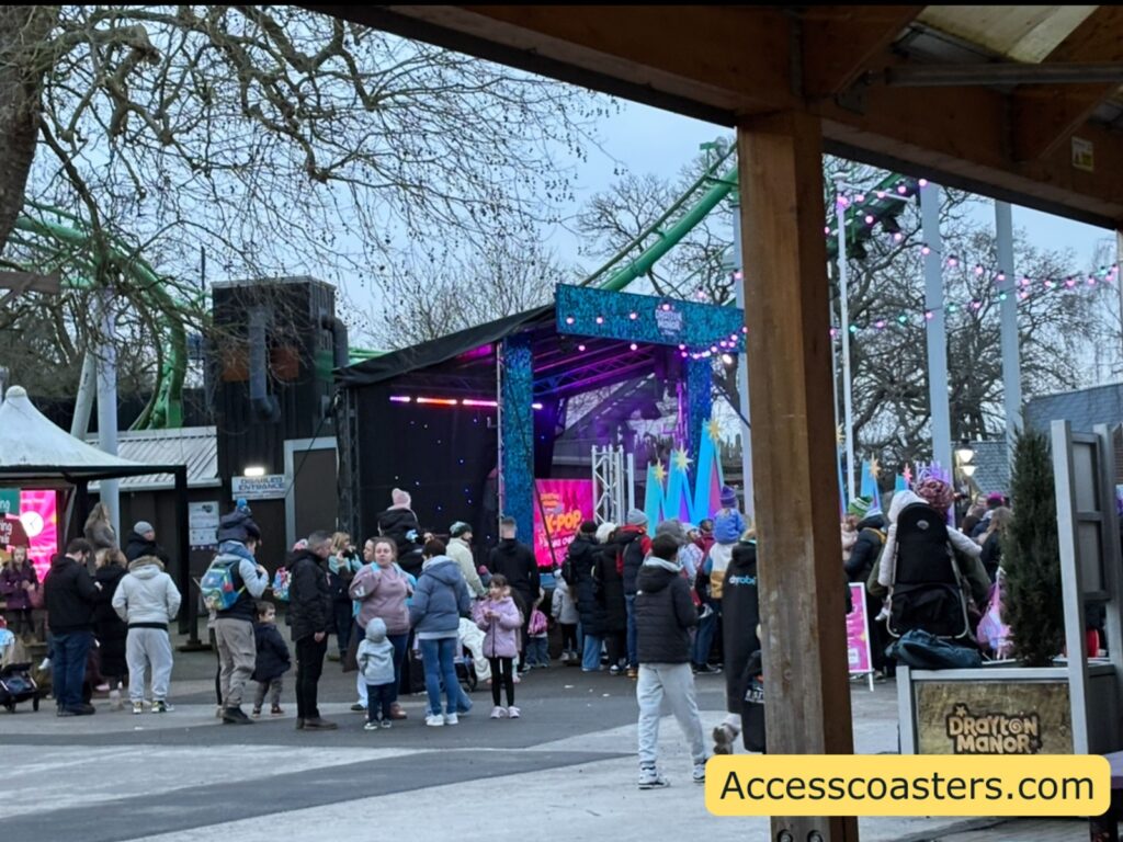 View of the K-pop concert from farther back near a covered picnic area, showing a busy crowd facing the stage through trees and support posts.