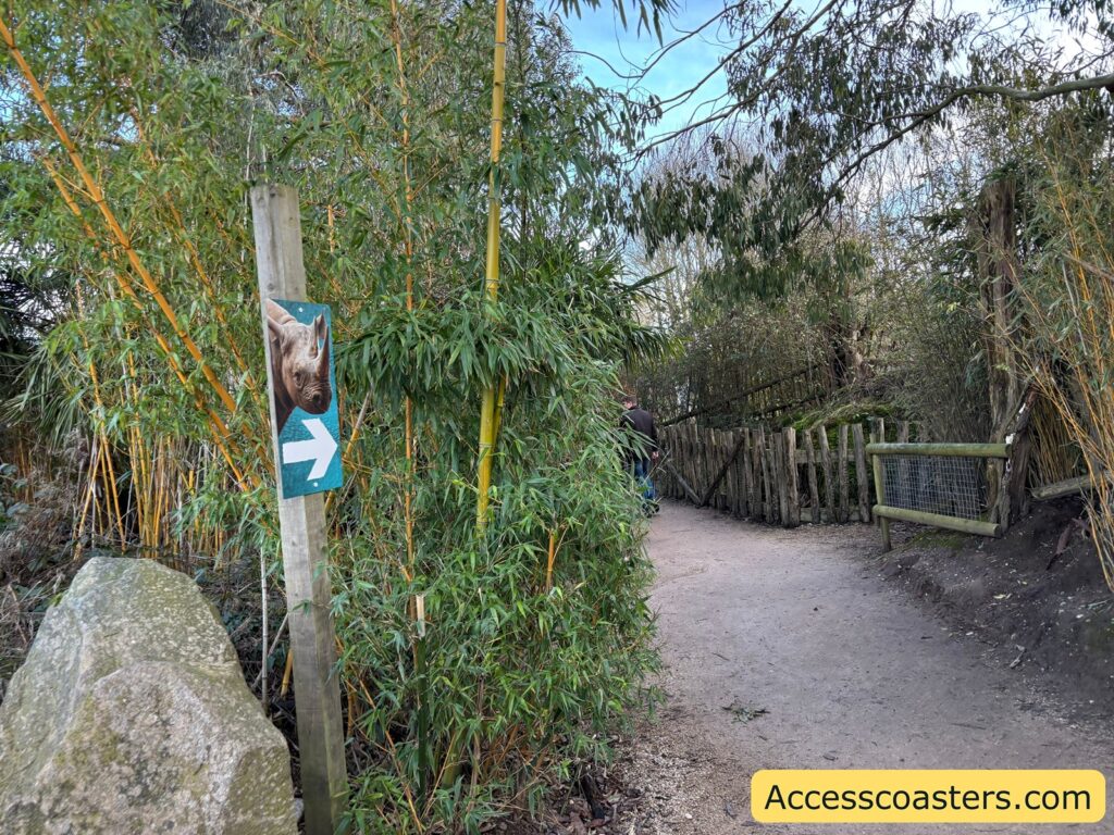 A gravel zoo path through dense greenery, with a sign showing a rhino and a right-pointing arrow toward the rhino area.
