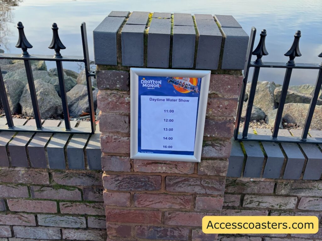 Brick pillar and black iron fence with a posted “Water Show Times” sign beside the lake.