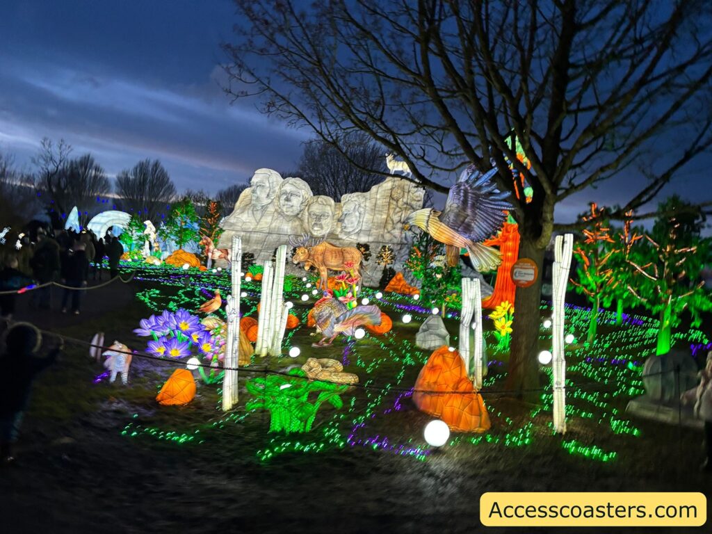 Night-time trail scene with green ground lights and multiple illuminated displays, including large pale mountain-like shapes and animal figures, with a leafless tree silhouetted overhead.