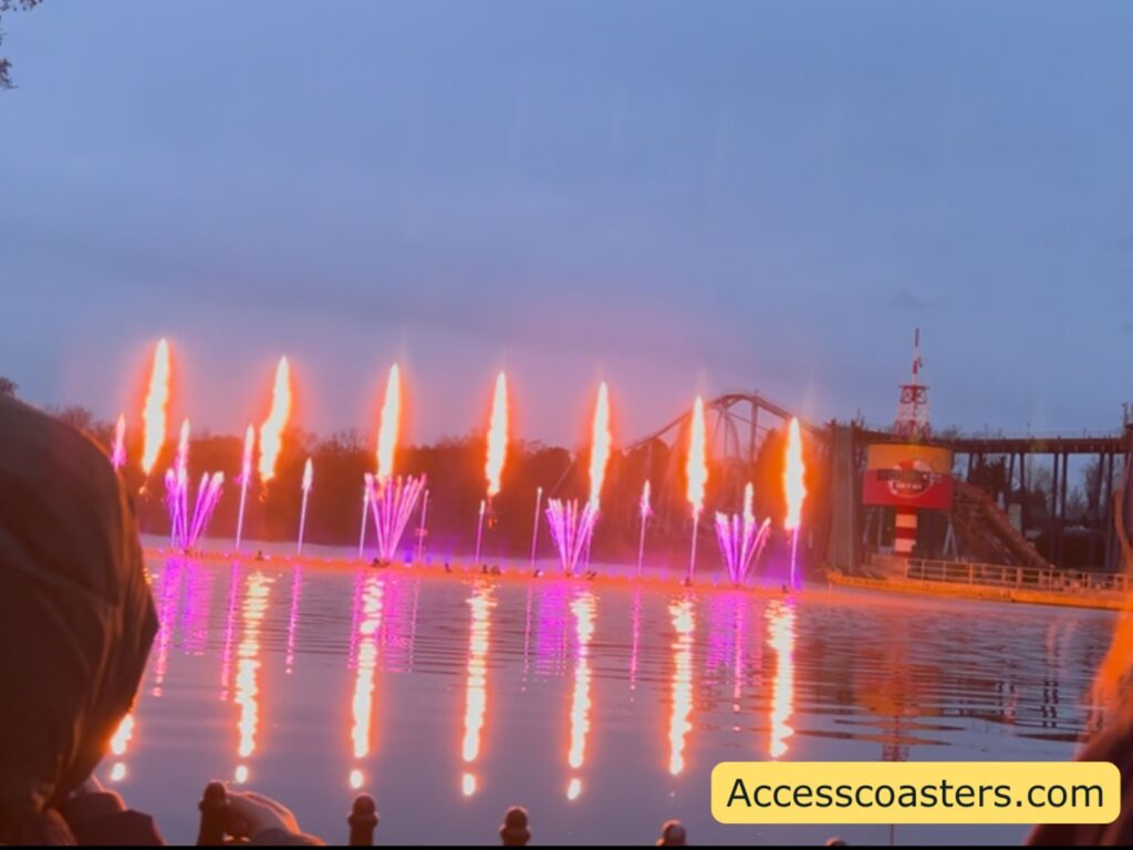 Dusk water show on a lake with pink and orange-lit fountains and bursts of flame, reflected on the water.