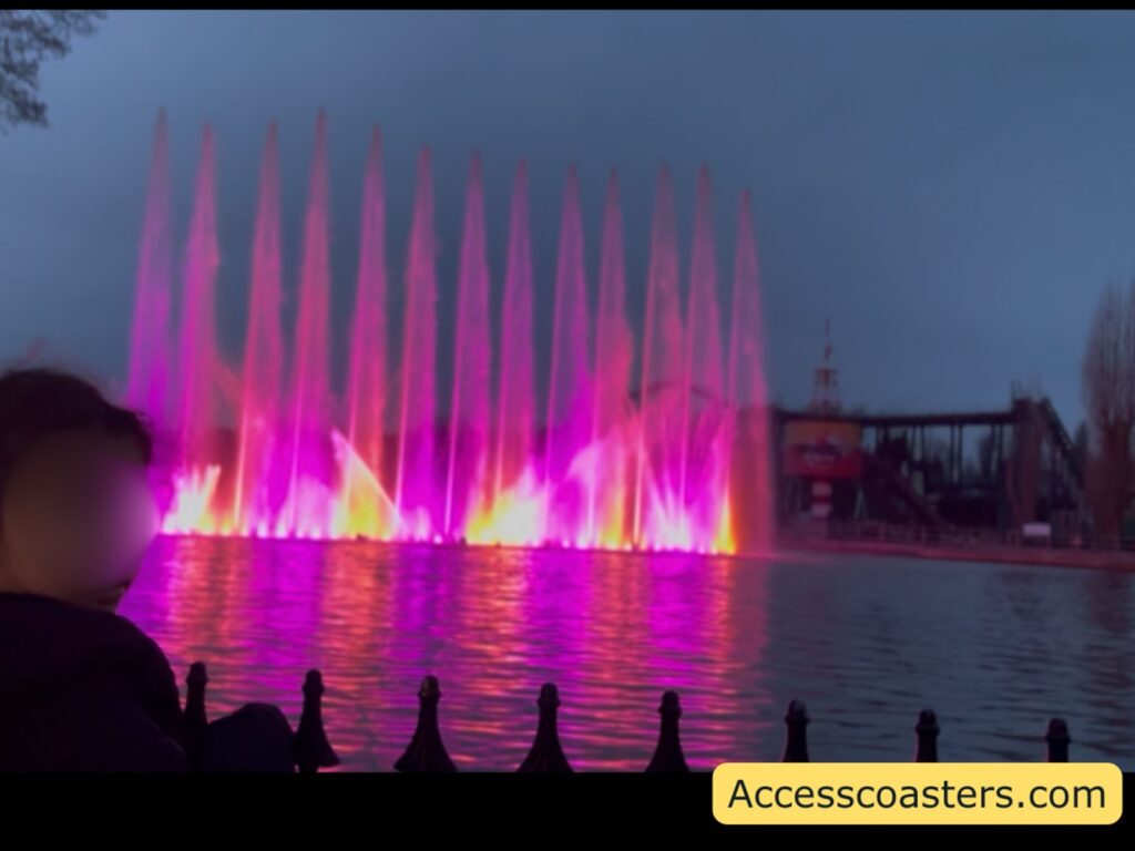 Tall pink-lit fountains rising in a row across the lake at dusk, reflected on the water.