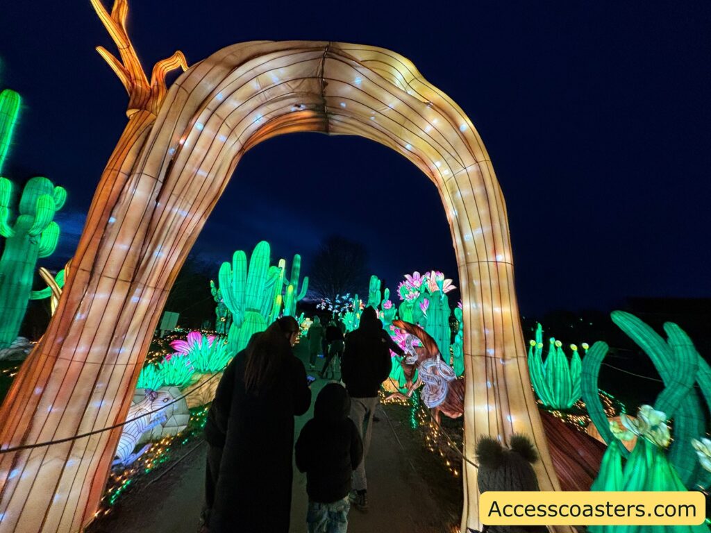 Nighttime light trail with a large glowing arch over the path, surrounded by illuminated green cactus sculptures and colorful flower lights, with several visitors walking through.