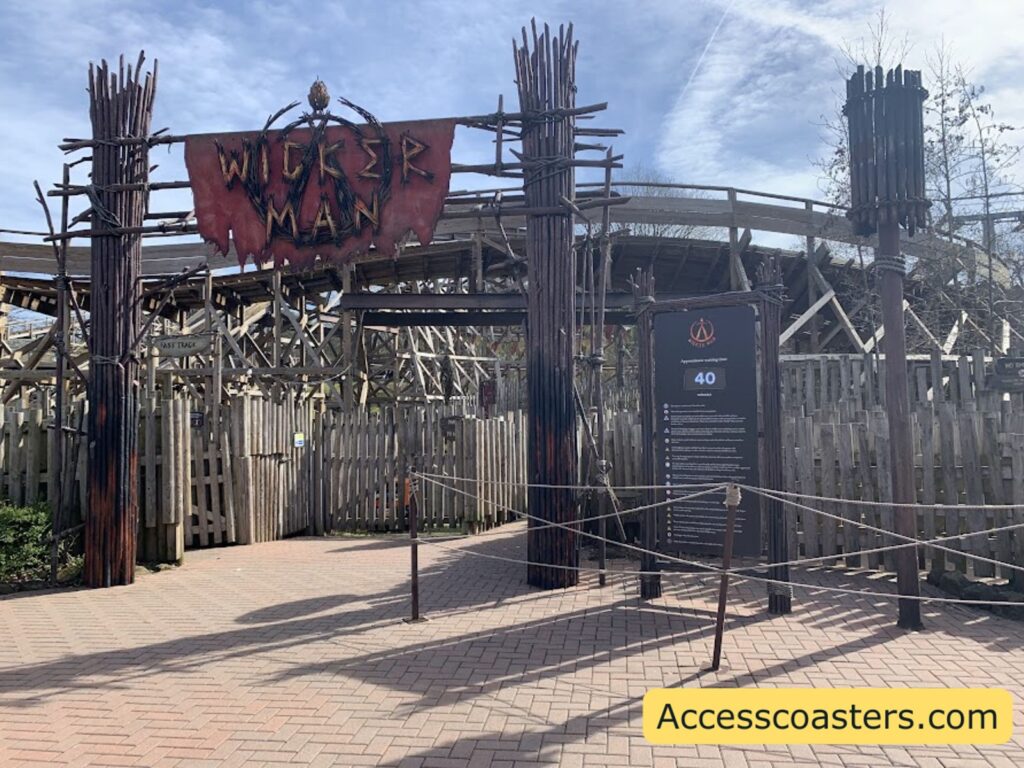 main entrance to the wicker man ride with ride logo above entrance as well as a digital queue time screen and ride restriction board.