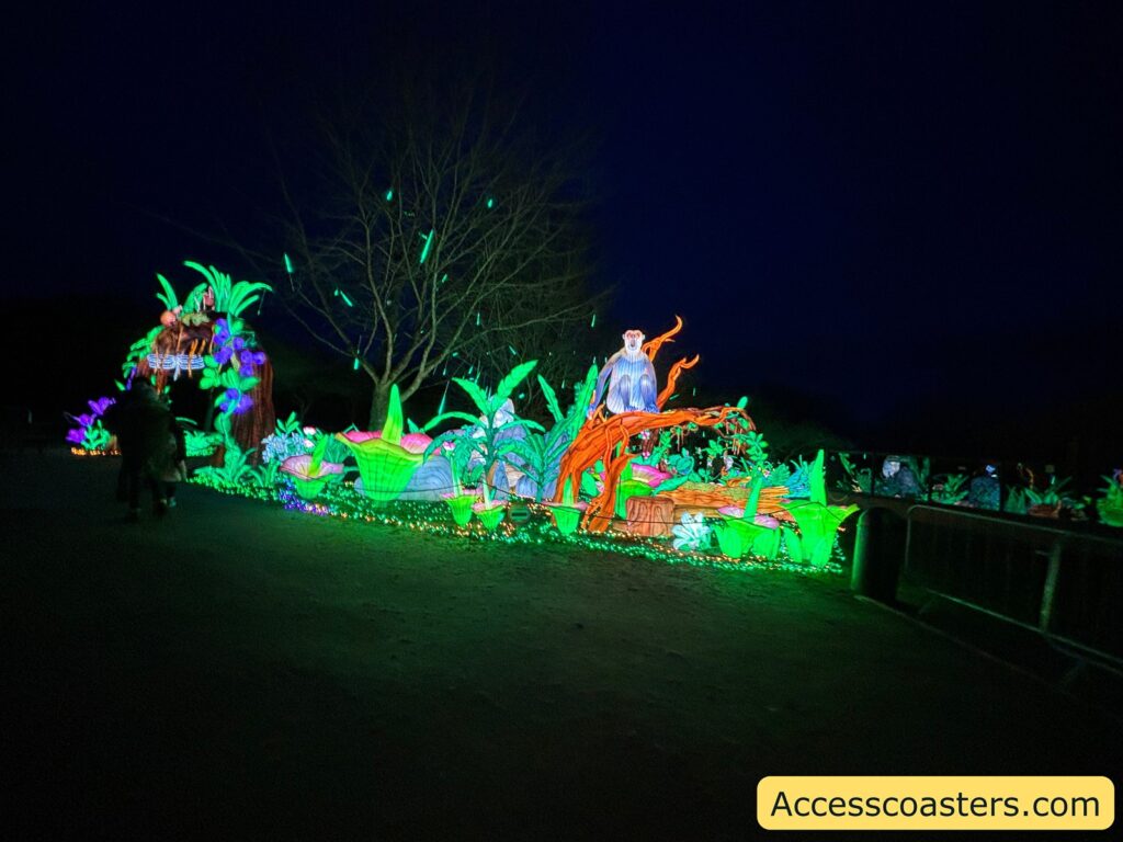 Nighttime photo of a light-trail walkway with colorful jungle-themed illuminated sculptures on the side, including tiger-like figures and glowing plants, with visitors walking past.