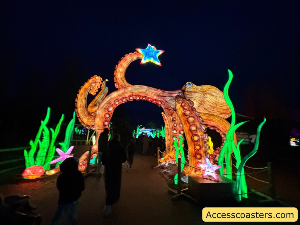Nighttime photo of an illuminated octopus archway forming an entrance to an underwater-themed light trail, with glowing green seaweed sculptures lining the path and visitors walking through.

