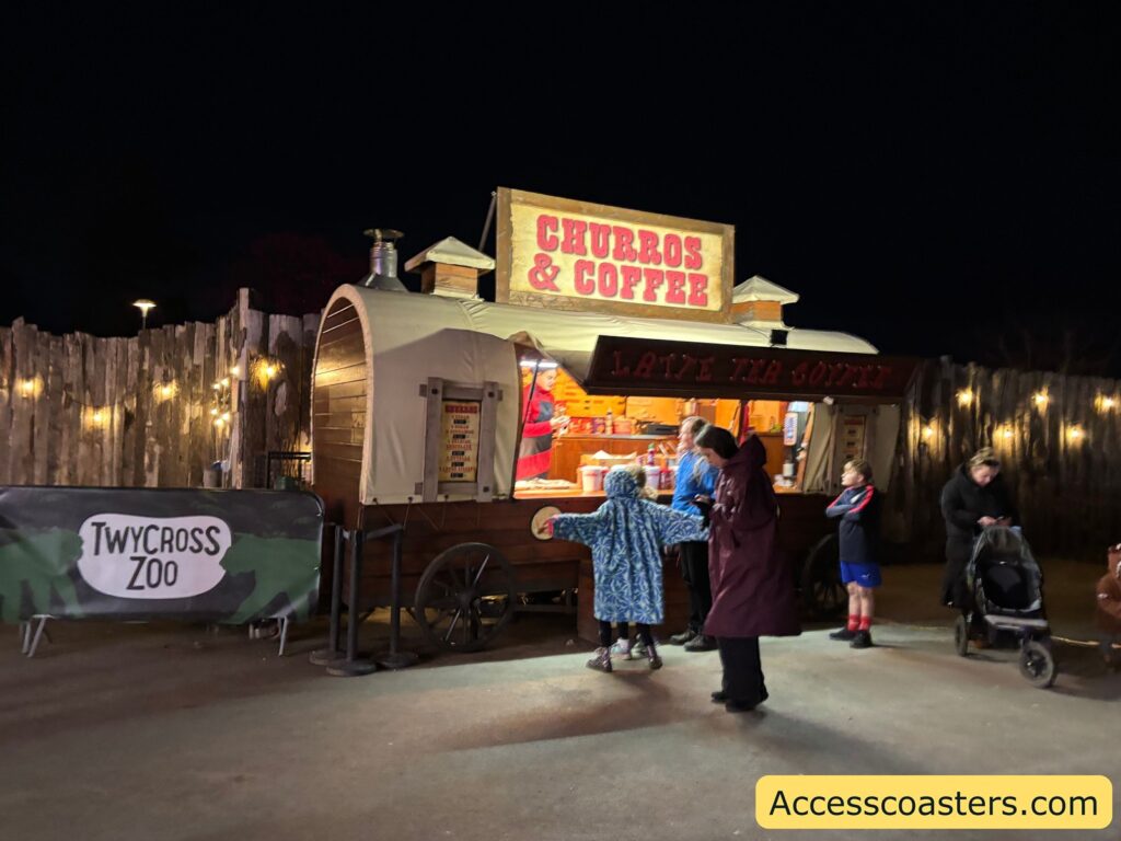 Closer nighttime view of the Food Village with a brightly lit “Churros & Coffee” stall and a “Twycross Zoo” sign, with visitors gathered at the counter.
