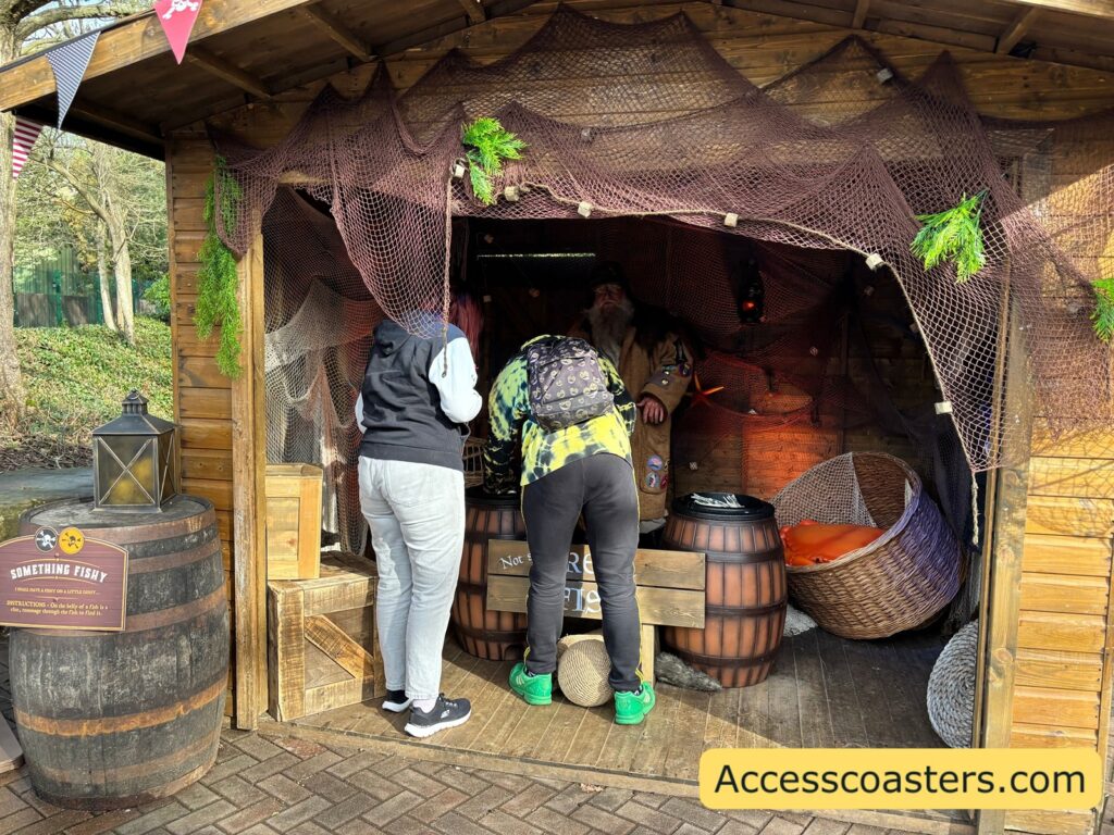 people standing at the entrance of a small wooden hut decorated with fishing nets and greenery; barrels sit around the doorway.