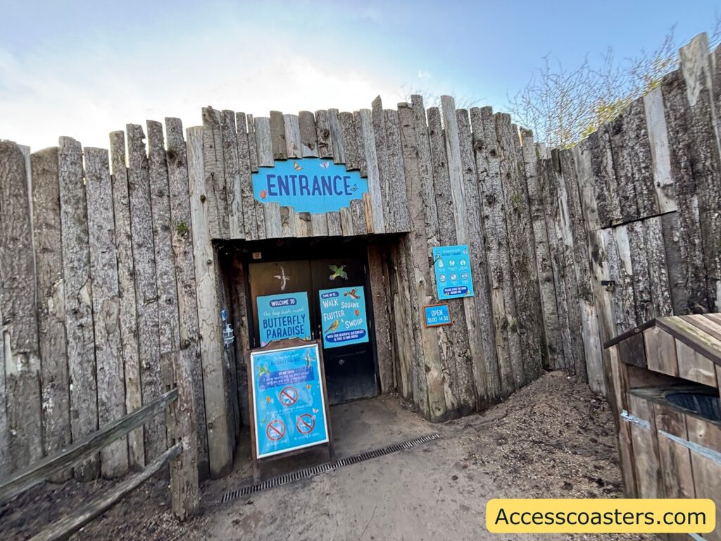 Outdoor attraction entrance made of vertical wooden fencing with an “Entrance” sign above a doorway and posters on the door.
