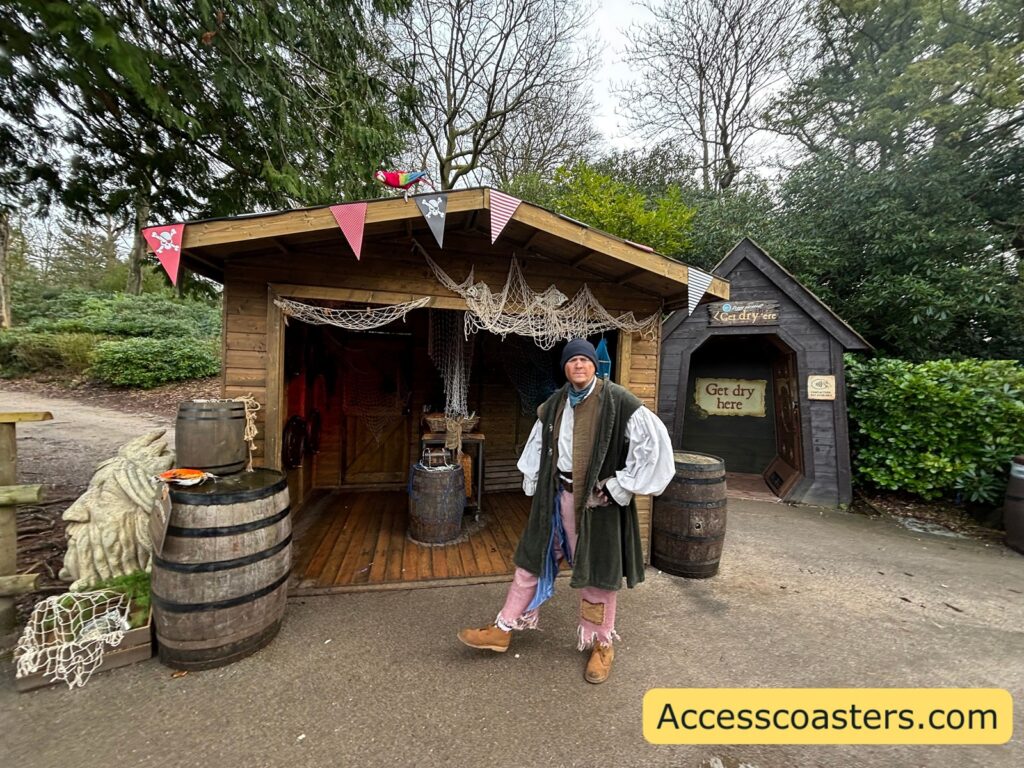 A person in pirate-like costume (hat/coat) standing in front of a small hut with bunting and barrels.