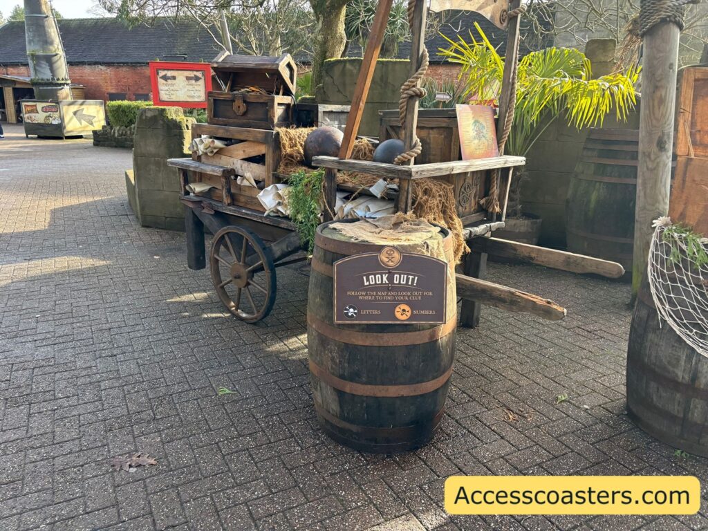 irate-style props on a paved area, including barrels and a wooden cart loaded with items; a sign is attached to a barrel with information about the letter and number trail
