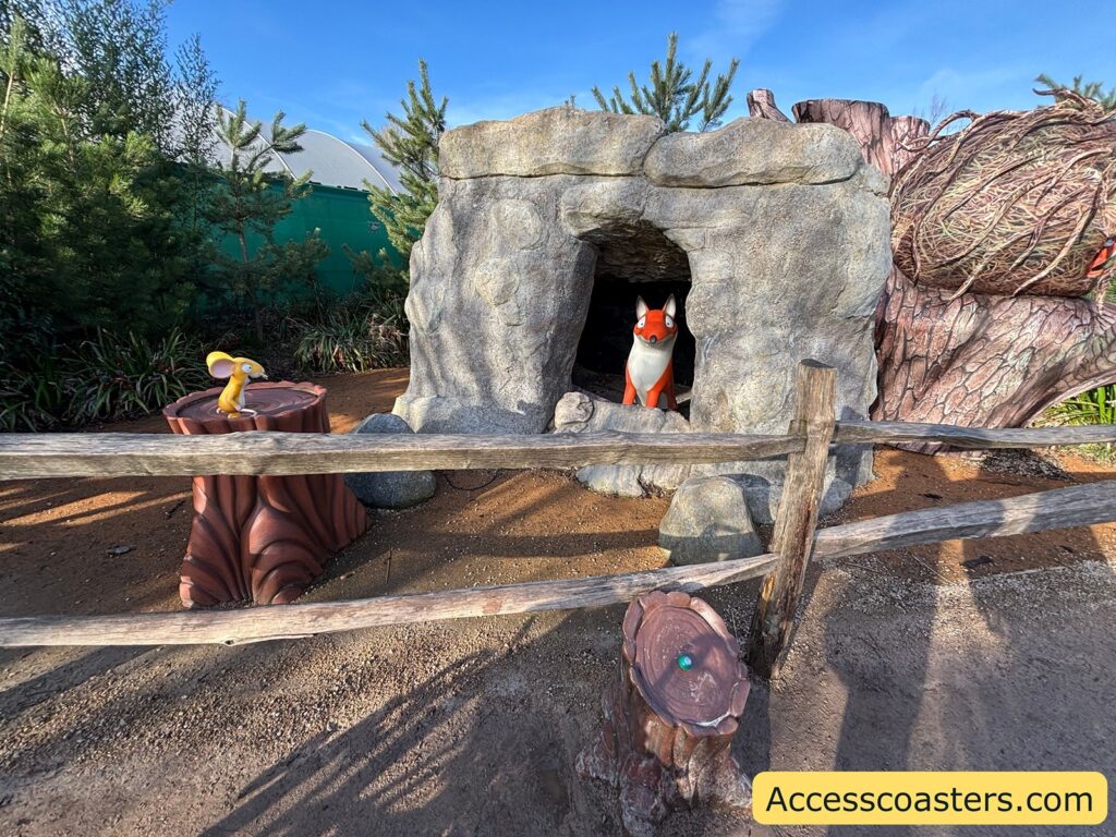 Fox-themed rock “house” with a round entrance opening behind a low wooden barrier; a fox figure is visible in the opening.

